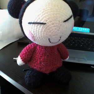 May include: A handmade crochet doll with a white head, black hair buns, and a red top. The doll has closed eyes, a small smile, and black legs. The doll is sitting on a dark surface.
