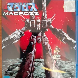 Puede incluir: Caja de disco Blu-ray de la serie de anime Macross, con una gran ilustración de robot en tonos grises y rojos. El título "MACROSS" se muestra de forma destacada, con caracteres japoneses encima. Una pequeña ilustración de una niña está a la derecha.