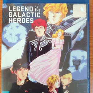 Puede incluir: Caja Blu-ray azul con el anime "Legend of the Galactic Heroes". La portada muestra personajes de anime en un entorno espacial. El título es blanco, con personajes con uniformes militares y una mujer con un vestido rosa.