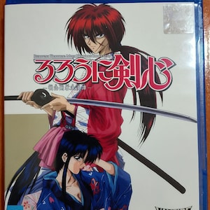 Puede incluir: Una caja Blu-ray azul con ilustraciones de anime. La portada muestra dos personajes, uno con cabello rojo empuñando una espada y otro con cabello negro con un kimono azul. Texto japonés visible. La caja está etiquetada como "Vampire House."
