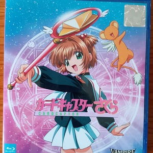 Puede incluir: Una caja azul de Blu-ray con ilustraciones de anime. La portada muestra a una chica con uniforme escolar sosteniendo un bastón y un personaje de oso de dibujos animados. El título "Cardcaptor Sakura" se muestra en japonés e inglés.