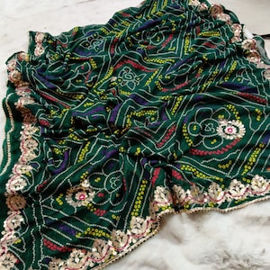 May include: A dark green textile with a detailed pattern of white, yellow, red, and purple dots and floral designs. The edges are trimmed with gold-colored embellishments. The fabric is draped on a white surface.