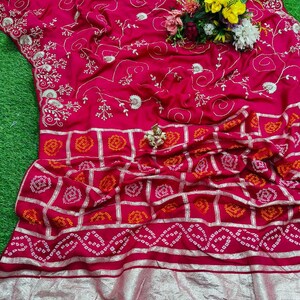 May include: A vibrant fuchsia saree with intricate gold embroidery and a patterned border. The fabric features delicate floral and geometric designs. A small bouquet of flowers rests on the saree.