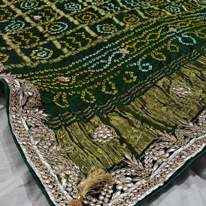 Puede incluir: Un textil verde oscuro con intrincados patrones dorados y blancos. La tela presenta un borde detallado con adornos plateados y una borla decorativa. El dise&ntilde;o general sugiere un estilo tradicional o &eacute;tnico.