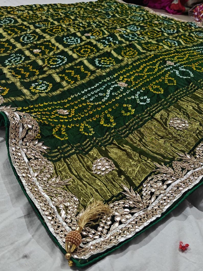 Puede incluir: Un textil verde oscuro con intrincados patrones dorados y blancos. La tela presenta un borde detallado con adornos plateados y una borla decorativa. El dise&ntilde;o general sugiere un estilo tradicional o &eacute;tnico.