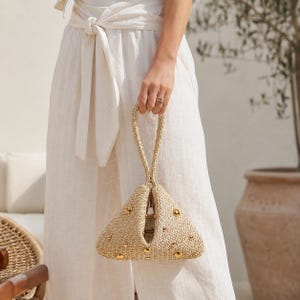 May include: A beige woven straw handbag with a triangular shape and a single top handle. The exterior is decorated with small, polished gold-tone metal studs scattered across the textured surface, creating a structured and compact accessory.