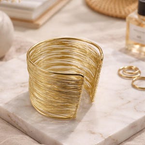 May include: A gold-colored cuff bracelet made of numerous thin wires. The open-ended bracelet sits on a white marble surface. The jewelry is a statement piece, suitable for adding a touch of elegance to any outfit.