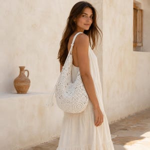 Può includere: Una borsa hobo all'uncinetto bianca caratterizzata da un intricato motivo floreale circolare e una chiusura a coulisse con estremità annodate. La borsa è realizzata in filato bianco e si indossa a spalla, mostrando una forma morbida.