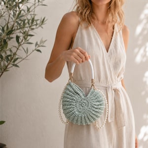 Pode incluir: Uma mala de crochet verde sálvia com um design canelado em semicírculo e um motivo circular central. A mala apresenta uma pega superior entrançada branca e uma alça decorativa de contas de pérolas brancas, fixadas com ferragens douradas.