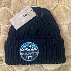 May include: Black knit beanie with a circular patch featuring a mountain range, blue sky, and the text "PATAGONIA 1973." A tag attached to the beanie reads "White w/Dolomite Blue 39045 WDOB SP18."