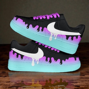 Puede incluir: Zapatillas negras con un logotipo Nike blanco y un diseño único. Las zapatillas presentan una parte superior negra, un degradado morado y azul en la entresuela y un efecto de pintura goteando. Los cordones son morados.