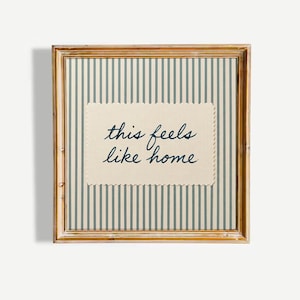 May include: Square framed artwork with a gold-coloured frame. The artwork features a cream-coloured rectangle with the phrase "this feels like home" in navy blue script. The background has vertical stripes in cream and light blue.
