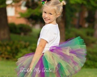 May include: A young girl wearing a green, gold, and purple tulle tutu skirt with a white shirt and brown cowboy boots. She is wearing a green, gold, and purple bow in her hair. The text "Tickle My Tutu" is visible on the skirt.