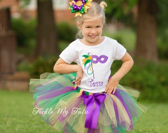 May include: A young girl wearing a white shirt with a Mardi Gras mask design and the name "Tessa" embroidered on it. She is also wearing a multi-layered tulle skirt in green, yellow, and purple with a purple satin bow. The skirt is labeled "Tickle My Tutu".