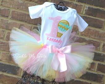 May include: A white onesie with a pink number one and a hot air balloon graphic. The onesie is personalized with the name "Kennedy". The onesie is paired with a rainbow tulle tutu with a pink satin bow.