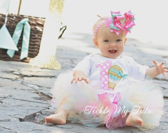 May include: A baby girl wearing a white onesie with a pink hot air balloon graphic and the number one on it. She is wearing a pink and turquoise bow headband and a white tulle tutu with pink and turquoise accents. The baby is sitting on a stone surface in front of a pink paper lantern and a gold number one.