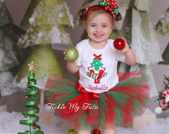 May include: A young girl wearing a white Christmas tree shirt with the name "Isabella" on it, a red and green tutu, and a red and white bow in her hair. She is holding a green and gold ornament in one hand and a red ornament in the other. She is standing in front of a backdrop of green and white Christmas trees.