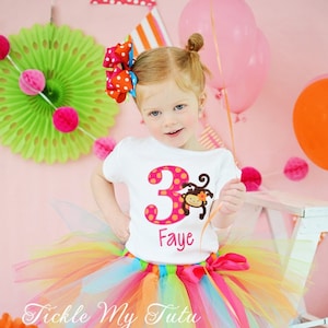 May include: A young girl wearing a white t-shirt with a pink number 3 and a monkey graphic. The shirt says "Faye" on the bottom. She is wearing a rainbow tulle tutu and teal shoes.