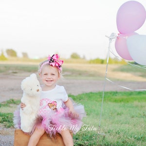 May include: A young girl wearing a pink tutu and a white shirt with a brown bear and the number two on it. She is holding a white teddy bear and sitting on a wooden crate. There are pink and white balloons in the background.