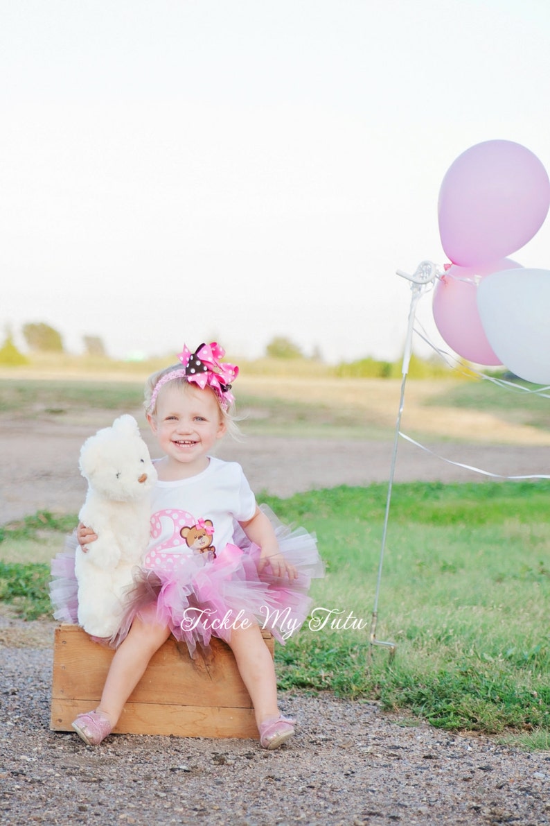 May include: A young girl wearing a pink tutu and a white shirt with a brown bear and the number two on it. She is holding a white teddy bear and sitting on a wooden crate. There are pink and white balloons in the background.