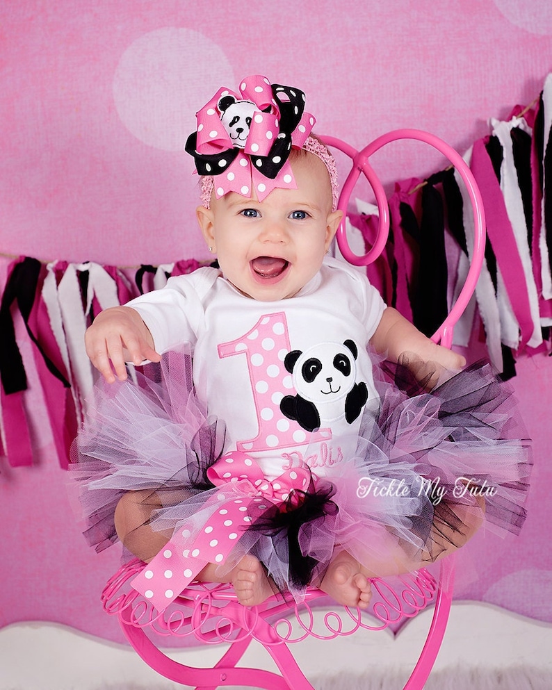 May include: A baby girl wearing a white onesie with a pink and white polka dot pattern and a large number one on the front. The onesie also has a black and white panda bear applique. The baby is wearing a black and pink tutu with a pink polka dot ribbon around the waist. The baby is wearing a pink and black polka dot headband with a panda bear on top. The baby is sitting on a pink metal chair. The text "Tickle My Tutu" is visible on the tutu.