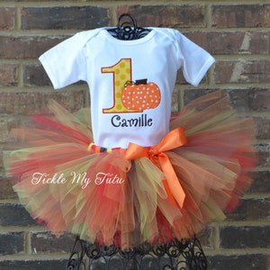 May include: A white onesie with a yellow number one with polka dots and a pumpkin on top. The onesie has the name "Camille" embroidered on it. The onesie is paired with a multi-layered tulle tutu in shades of orange, yellow, and brown.