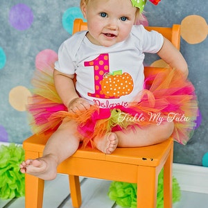 May include: A baby girl wearing a white onesie with a pink and white polka dot number one and a pumpkin design. The onesie says "Delanie" and the baby is wearing a multi-colored tulle tutu. The baby is sitting on a bright orange chair in front of a gray background with colorful polka dots.