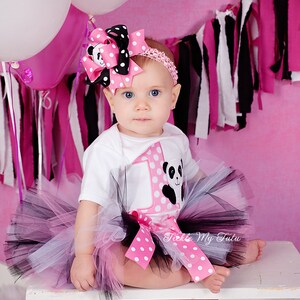 May include: A baby girl wearing a white onesie with a pink and white polka dot design and a panda bear graphic. The onesie has a large number "1" on the front. The baby is also wearing a black and pink tutu with a pink and white polka dot bow. The baby is wearing a pink and black polka dot headband with a bow. The background is pink with black and white streamers.