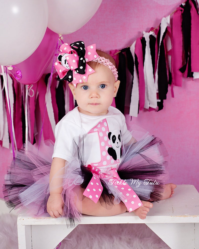 May include: A baby girl wearing a white onesie with a pink and white polka dot design and a panda bear graphic. The onesie has a large number "1" on the front. The baby is also wearing a black and pink tutu with a pink and white polka dot bow. The baby is wearing a pink and black polka dot headband with a bow. The background is pink with black and white streamers.