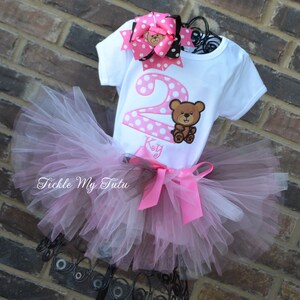 May include: A pink and gray tulle tutu with a pink satin bow. A white t-shirt with pink polka dots and a brown bear applique. The shirt says "2" and "Kay" on it. A pink and black bow with a brown bear applique.
