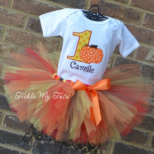 May include: A white onesie with a yellow number one and a pumpkin design. The onesie has the name "Camille" embroidered on it. The onesie is paired with a multi-layered tulle skirt in shades of orange, yellow, and red.