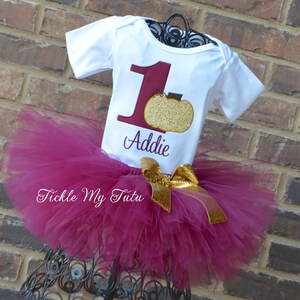 May include: A white onesie with a burgundy number one and a gold glitter pumpkin. The onesie is personalized with the name "Addie". The onesie is paired with a burgundy tulle tutu with a gold bow.