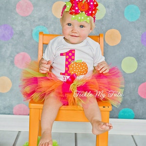 May include: A baby girl wearing a white onesie with a large orange pumpkin and the number one on it. She is wearing a pink and orange tulle tutu and a pink and green polka dot bow. She is sitting on a bright orange chair in front of a gray background with colorful polka dots.