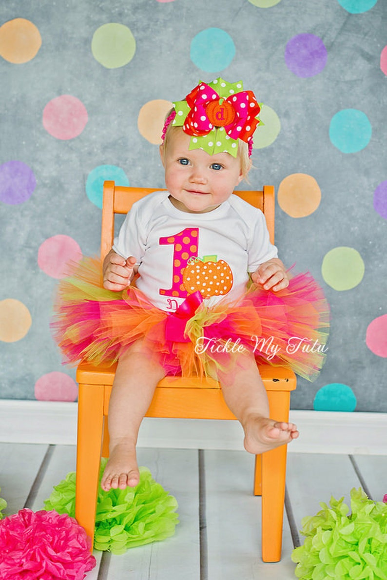 May include: A baby girl wearing a white onesie with a large orange pumpkin and the number one on it. She is wearing a pink and orange tulle tutu and a pink and green polka dot bow. She is sitting on a bright orange chair in front of a gray background with colorful polka dots.