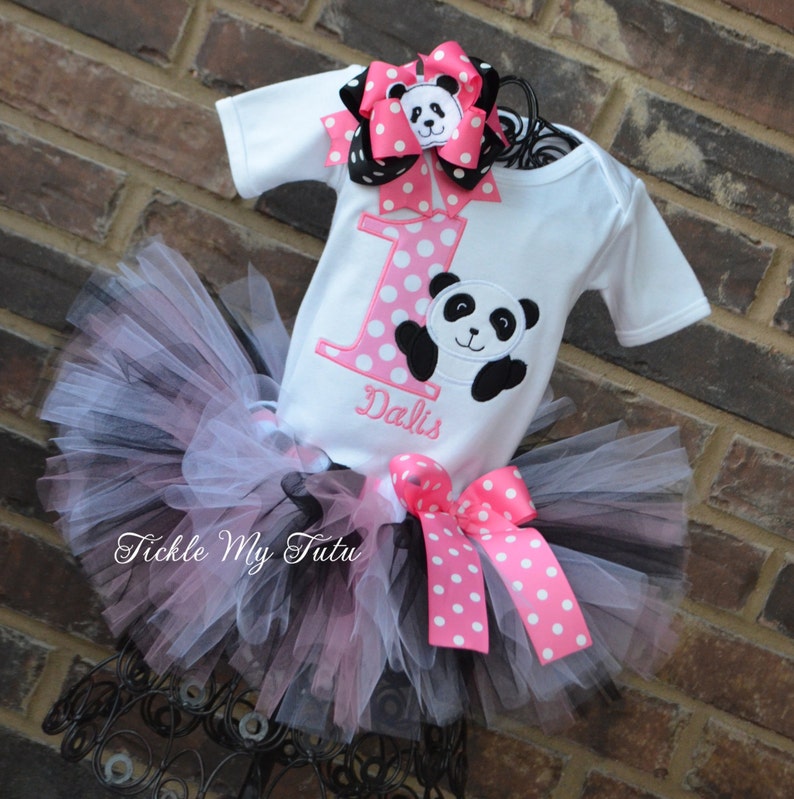 May include: A white onesie with a pink and white polka dot number one and a black and white panda bear. The onesie is personalized with the name "Dalis". The onesie is paired with a black and pink tulle tutu with a pink and white polka dot bow.