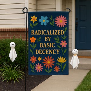 Op de afbeelding: Een marineblauwe tuinvaandel met de tekst "RADICALIZED BY BASIC DECENCY" in gouden letters, omringd door kleurrijke bloemmotieven. Twee kleine spookdecoraties staan aan weerszijden van de vlag.