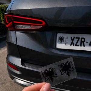 May include: Two black eagle decals are held in front of a grey car's rear. The decals feature a double-headed eagle design. The car has a license plate with the text "QLD XZR 4".