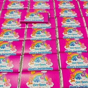 May include: A collection of rectangular chocolate bars wrapped in bright pink paper featuring a Care Bears graphic with a rainbow, clouds, and the text Hazel's 2nd birthday. Each wrapper displays the same colourful cartoon design and birthday message.