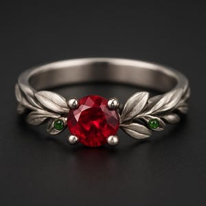 Puede incluir: Un anillo plateado con una gran gema roja y redonda como punto focal. La banda del anillo está adornada con detalles en forma de hojas, y pequeñas gemas verdes están engastadas a ambos lados de la piedra central. El anillo está sobre un fondo oscuro.