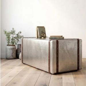 Puede incluir: Una mesa de centro rectangular plateada met&aacute;lica con un dise&ntilde;o remachado y correas de cuero marr&oacute;n. La mesa tiene esquinas redondeadas y est&aacute; coronada con libros y una br&uacute;jula. La mesa mide 122 cm de largo, 61 cm de ancho y 46 cm de alto.