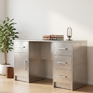 May include: A silver metal desk with a riveted design. The desk has a door and drawers with silver handles. A small mirror and a stack of books sit on the desk. A potted plant and a wooden container are in the background.