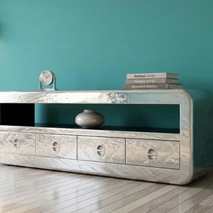 Puede incluir: Un mueble de televisión rectangular hecho de aluminio pulido con acabado remachado. Cuenta con cuatro cajones con tiradores metálicos circulares, un estante de almacenamiento abierto y una superficie superior lisa con un reloj y tres libros.