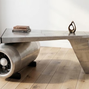 May include: An industrial coffee table with a riveted metal top and a jet engine base. The table has a bronze sculpture and two books on top. The table is approximately 152 cm long. A unique piece for a living room.