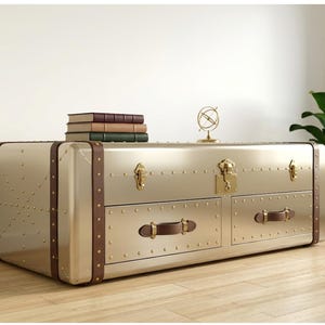 Puede incluir: Una mesa de centro dorada con forma de ba&uacute;l, con detalles en cuero marr&oacute;n y herrajes de lat&oacute;n. La mesa tiene dos cajones con tiradores de cuero y remaches decorativos. Libros y un peque&ntilde;o adorno de globo encima.