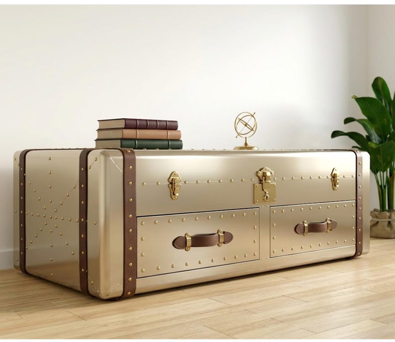 Puede incluir: Una mesa de centro dorada con forma de ba&uacute;l, con detalles en cuero marr&oacute;n y herrajes de lat&oacute;n. La mesa tiene dos cajones con tiradores de cuero y remaches decorativos. Libros y un peque&ntilde;o adorno de globo encima.
