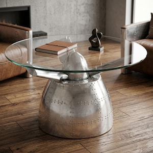 Könnte beinhalten: Ein einzigartiger Couchtisch mit einer transparenten Glasplatte und einem vernieteten, polierten Aluminiumsockel in Form eines Flugzeugpropellers. Der Tisch hat eine runde Glasplatte. Eine kleine Skulptur und ein Notizbuch liegen auf dem Tisch.