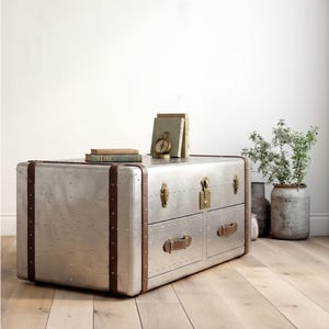 Puede incluir: Un mueble tipo ba&uacute;l de metal plateado con correas y asas de cuero marr&oacute;n. El mueble tiene dos cajones y est&aacute; adornado con remaches. Libros y una br&uacute;jula encima. Un mueble decorativo.