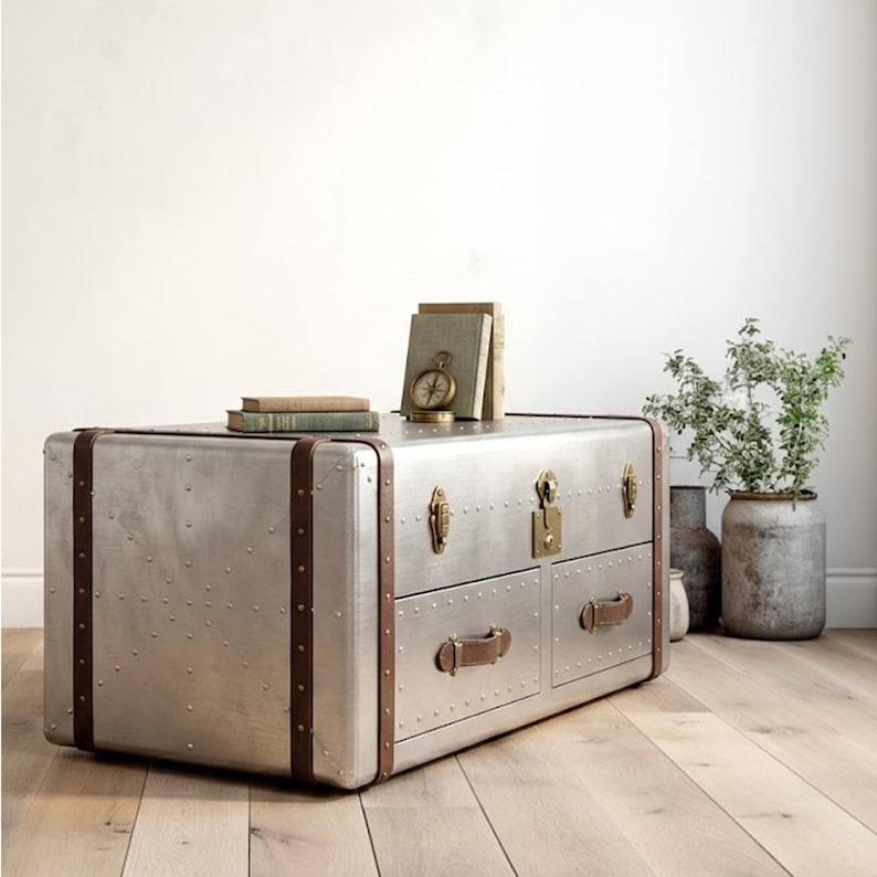 Puede incluir: Un mueble tipo ba&uacute;l de metal plateado con correas y asas de cuero marr&oacute;n. El mueble tiene dos cajones y est&aacute; adornado con remaches. Libros y una br&uacute;jula encima. Un mueble decorativo.
