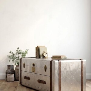Puede incluir: Un mueble de metal plateado estilo ba&uacute;l con dos cajones, detalles en cuero marr&oacute;n y herrajes de lat&oacute;n. El mueble tiene un dise&ntilde;o remachado y est&aacute; coronado con libros y un peque&ntilde;o reloj. El mueble est&aacute; sobre un suelo de madera.