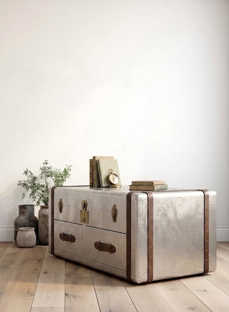 Puede incluir: Un mueble de metal plateado estilo ba&uacute;l con dos cajones, detalles en cuero marr&oacute;n y herrajes de lat&oacute;n. El mueble tiene un dise&ntilde;o remachado y est&aacute; coronado con libros y un peque&ntilde;o reloj. El mueble est&aacute; sobre un suelo de madera.